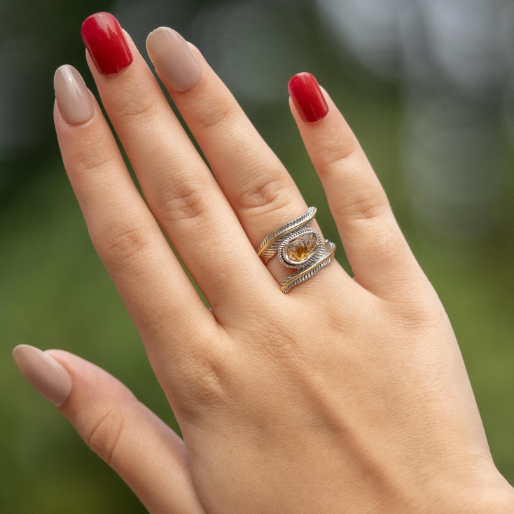 Hand wearing a silver ring with a yellow gemstone against a blurred green background